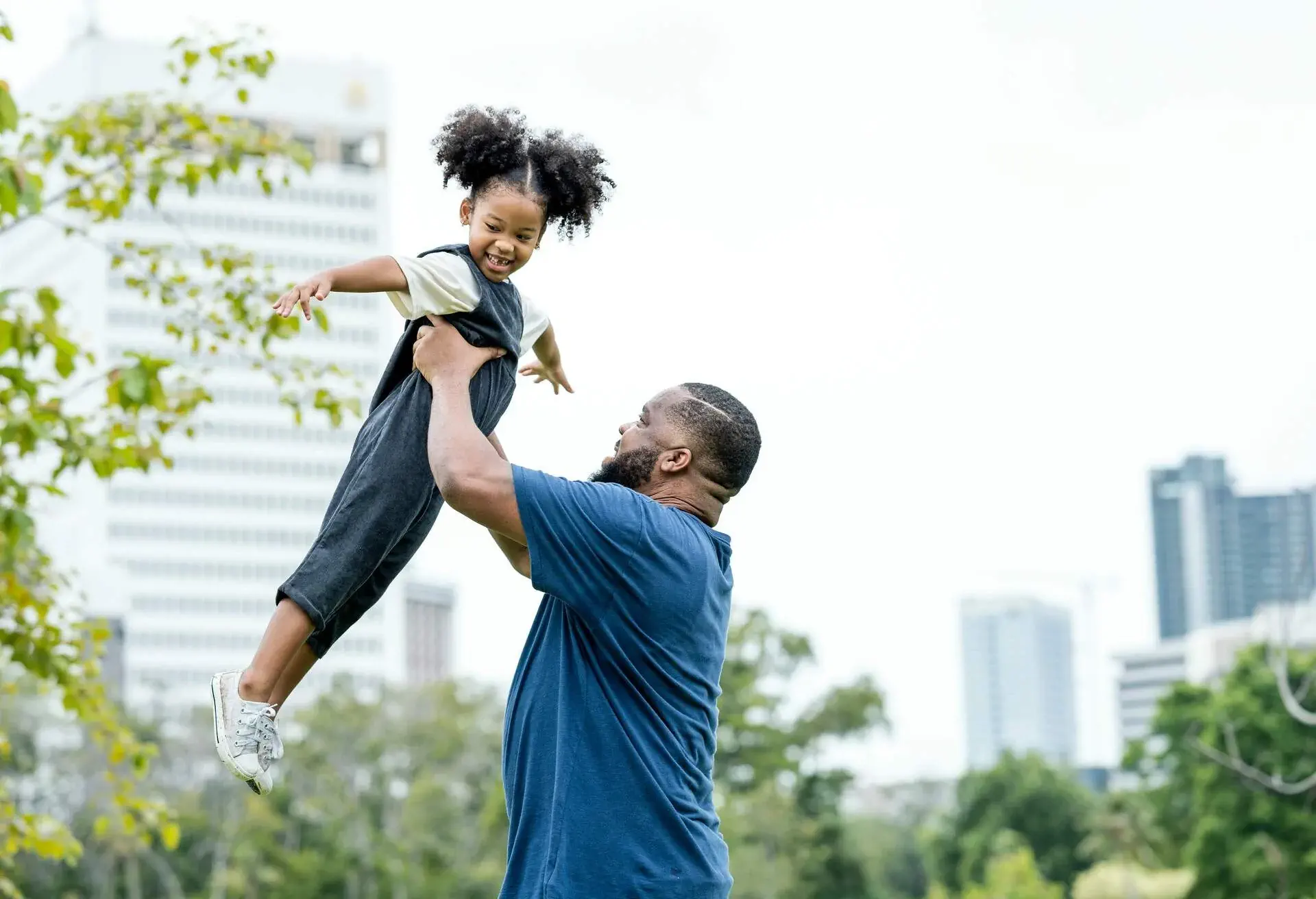 Playful Father carrying, picking up his daughter against skyscraper at public park.