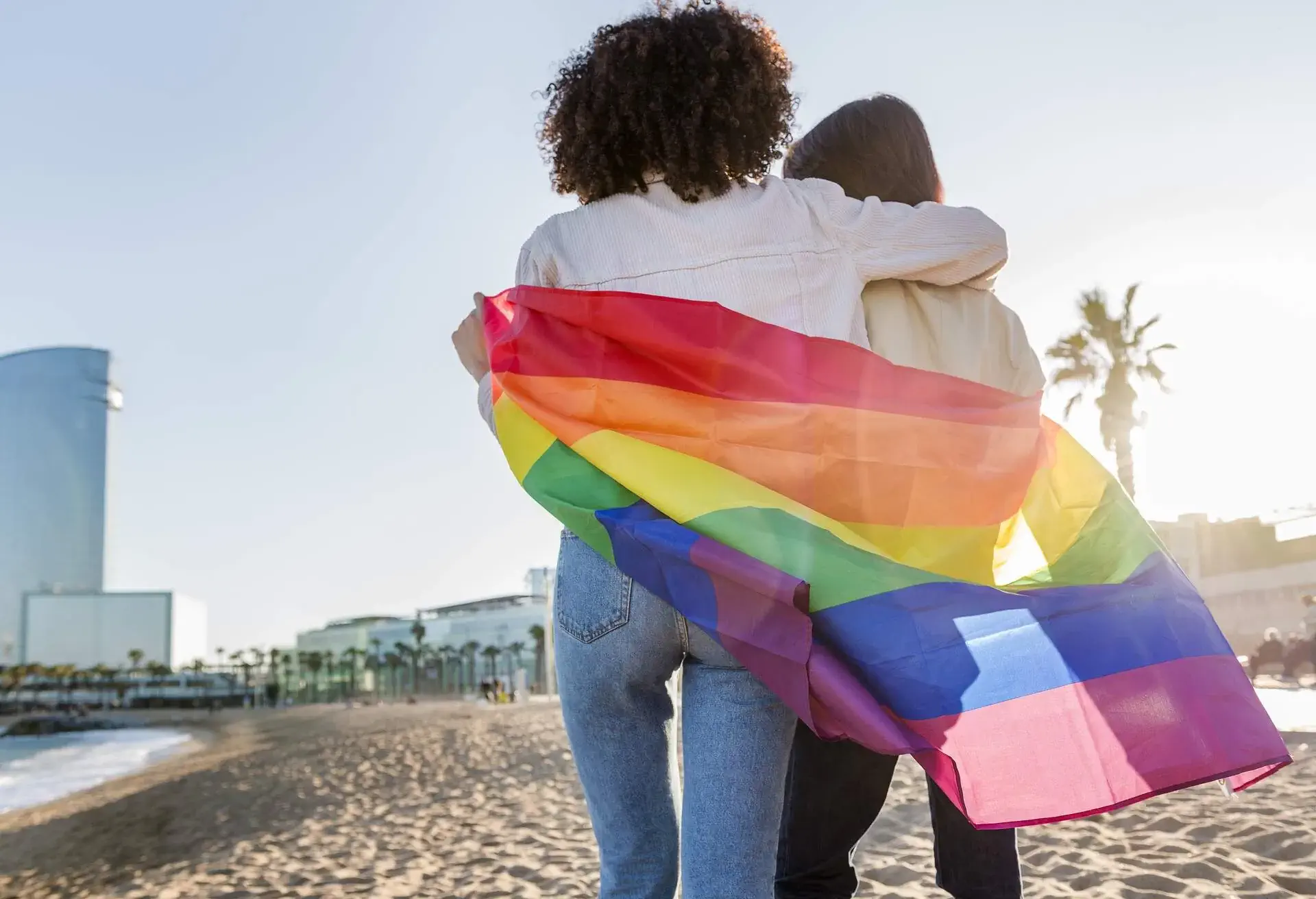 interracial couple of gay girls walking in front of the sea at Barcelona wrapped in the rainbow flag, symbol of the struggle for homosexual rights, concept of sexual freedom and racial diversity