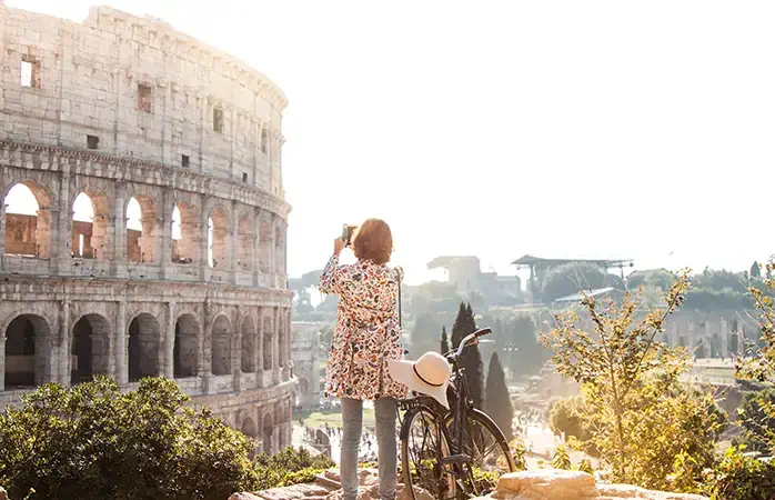 Beautiful young woman in colorful fashion dress alone on a hill with bike takes pictures of colosseum in Rome using smartphone camera at sunset. Attractive tourist girl with elegant straw hat.