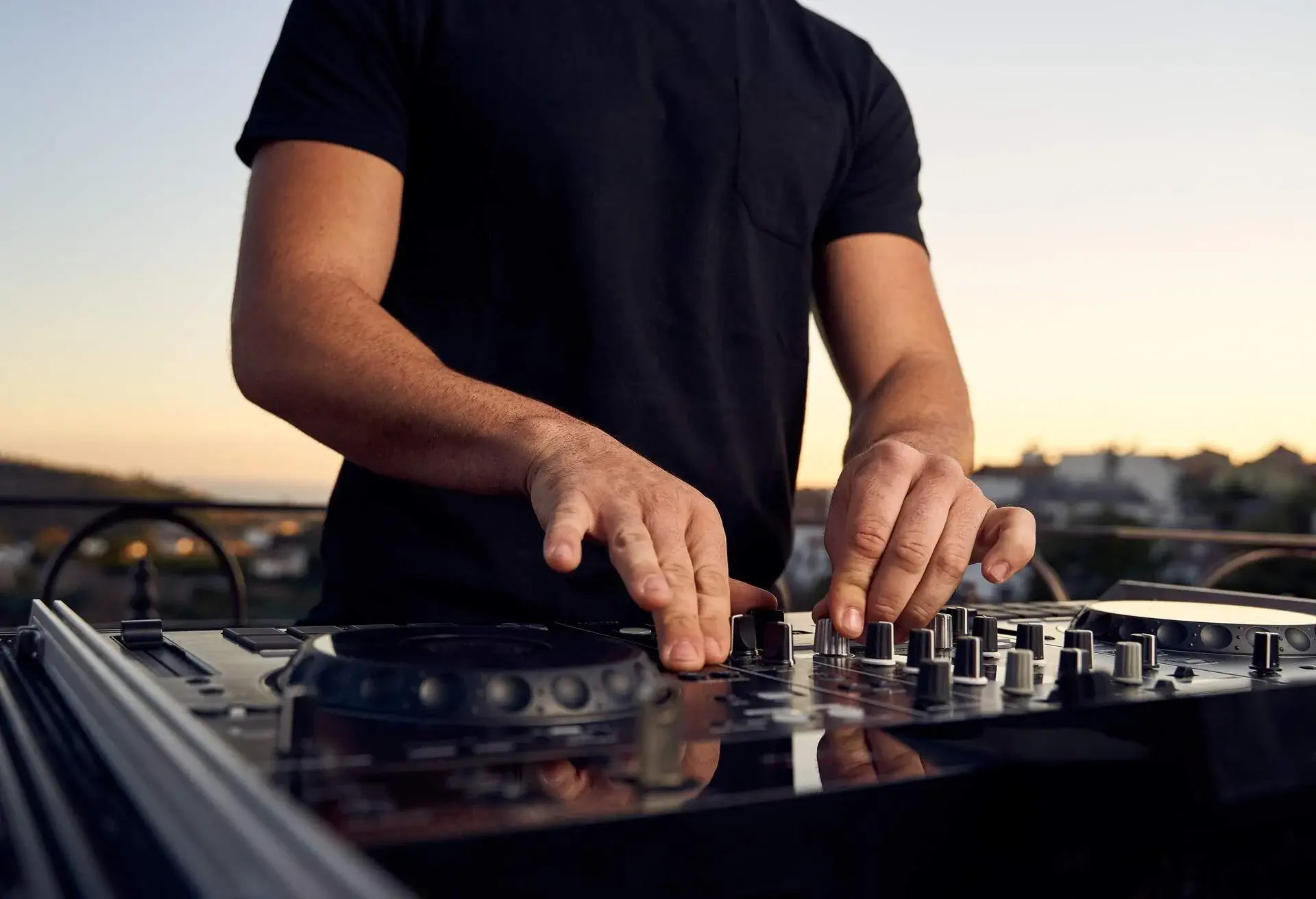 Unrecognizable male DJ hands playing music in a session with a mixer at a sunset party on a terrace. Electronic music concept.