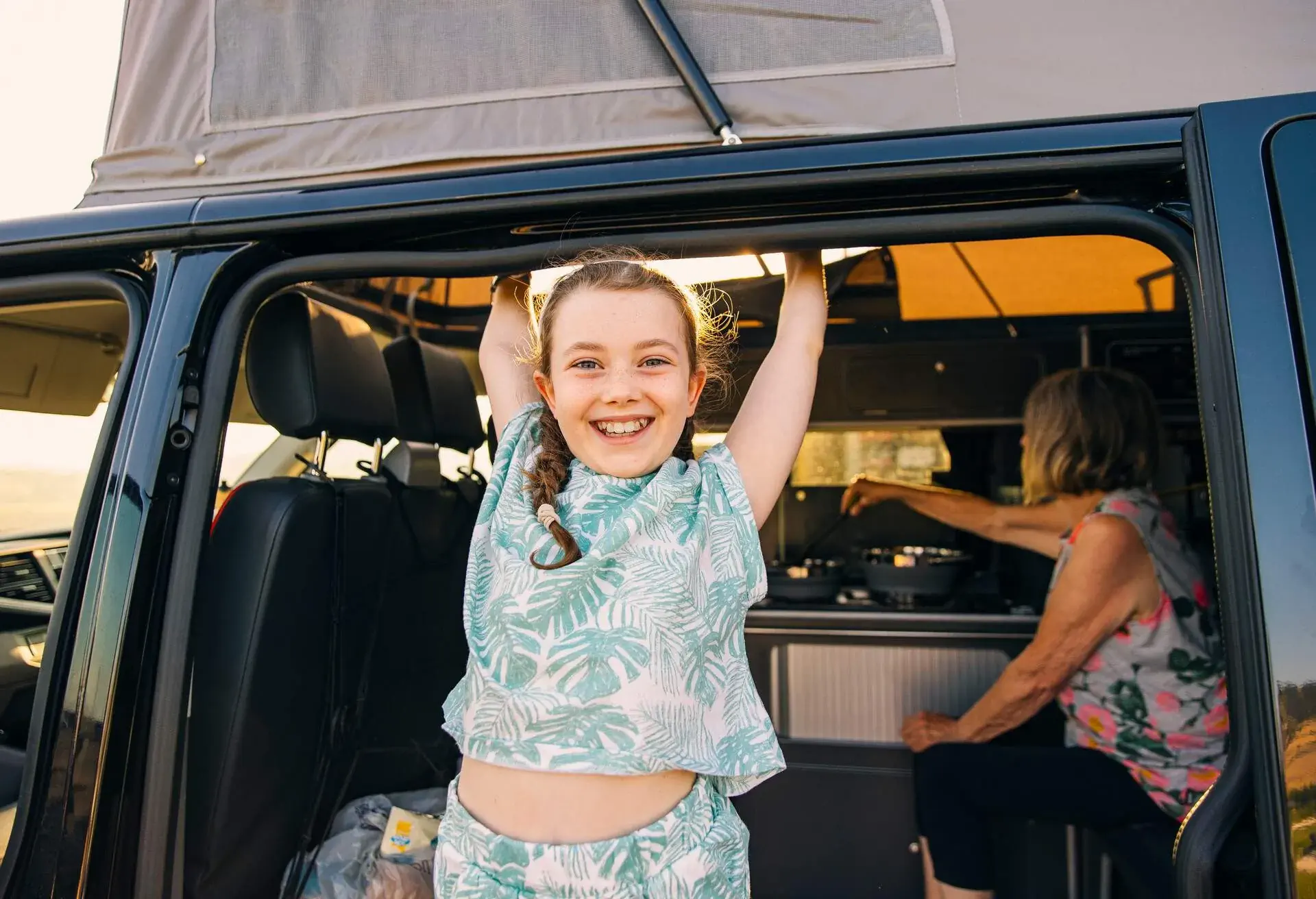 A happy young girl smiling from her camper van. In the background, her grandmother is preparing dinner.