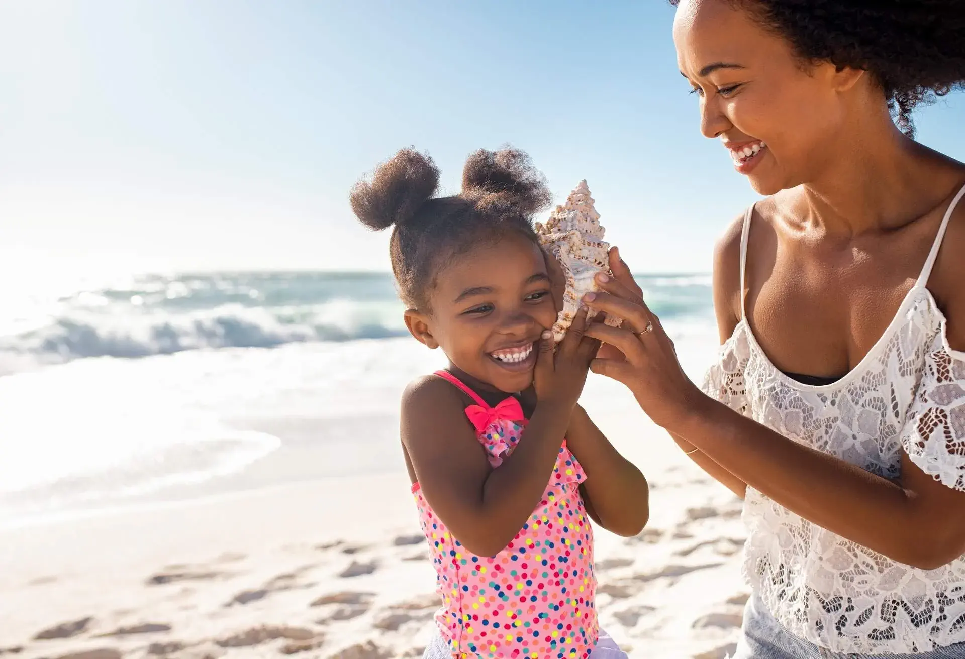 Beautiful young mother holding seashell near happy daughter ear at beach while she listening the sound of the ocean. Cute little female child hearing to cockleshell with mom at tropical beach. Laughing little child with woman listening to a shell at the beach.