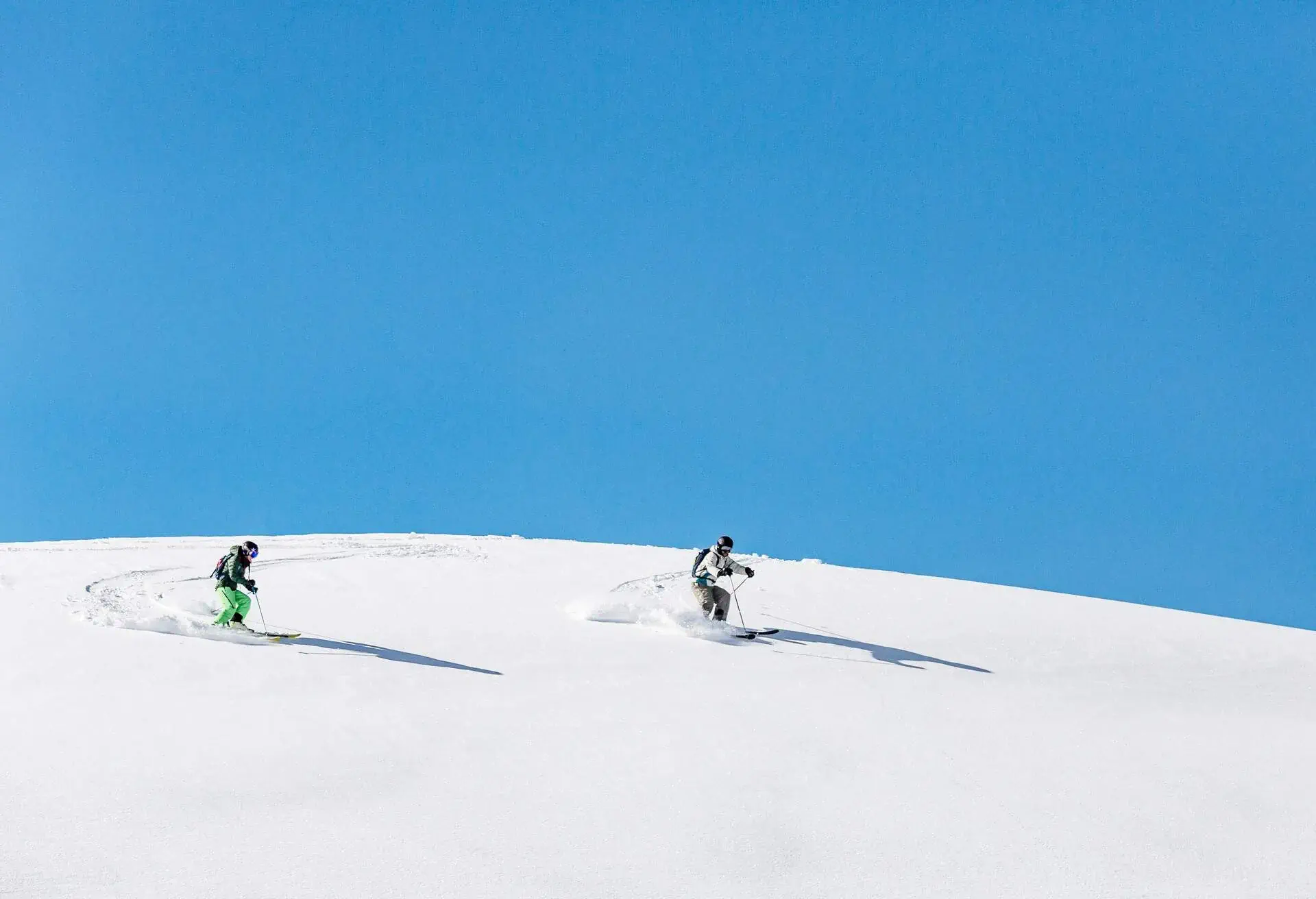 Two people skiing down an off-piste slope covered in fresh snow under the blue sky.