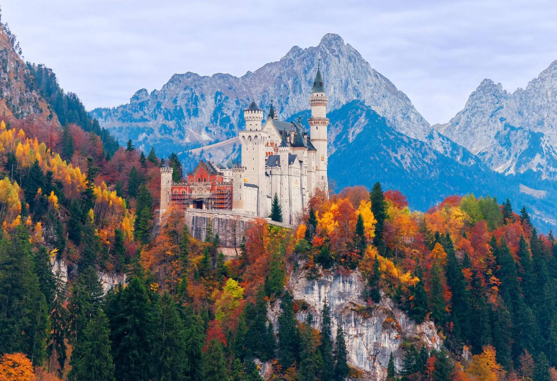 A medieval white castle with blue turrets on high alpine foothills surrounded by tall trees in autumn foliage and a snow-capped mountain range in the background.