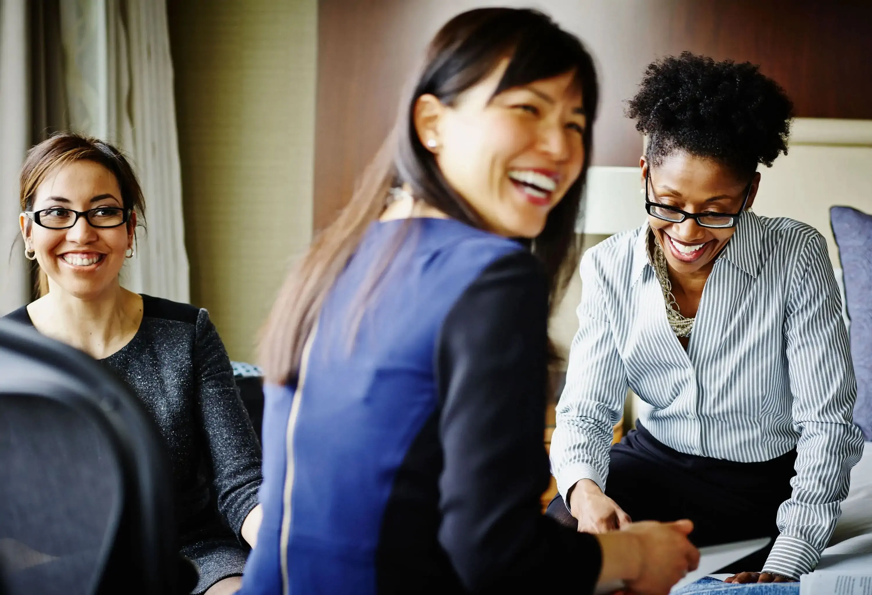 Three women in business attire smile while sitting on a bed.